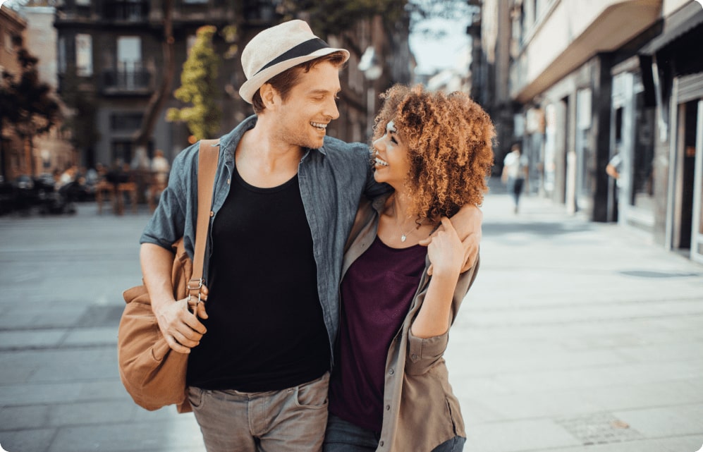 A young white male with hat looks affectionately into the eyes of his black female partner while walking through their college campus